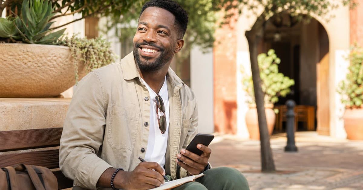 A smiling professional sits outdoors with a phone and notebook, pausing with a pen over the page in warm daylight.
