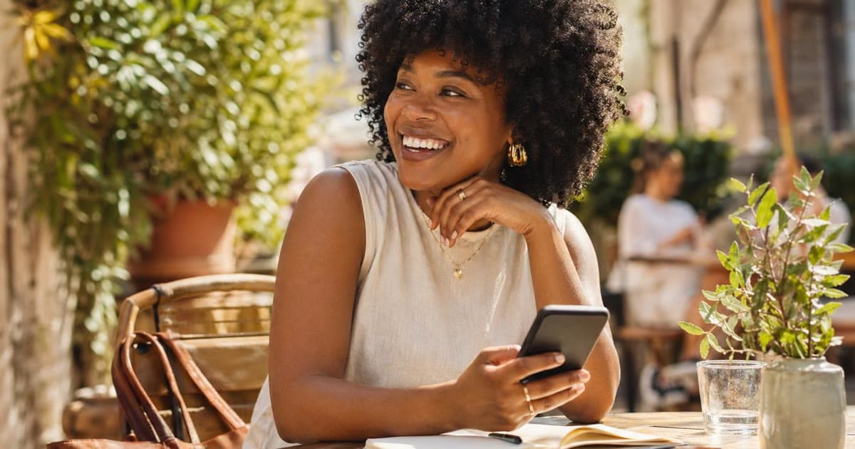 A smiling Black woman sits at a sunny outdoor cafe holding a phone above an open notebook beside a glass of water.