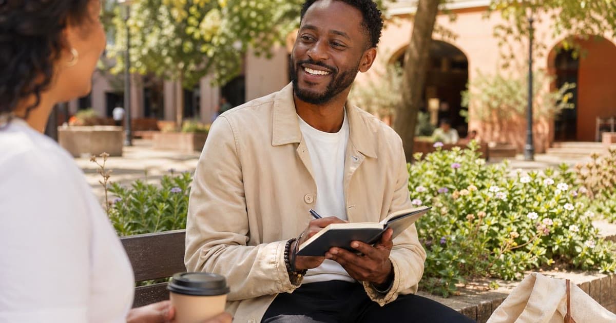 A Black man writes in an open notebook while talking with a woman on a sunny garden bench beside one takeaway coffee cup and one ceramic mug.
