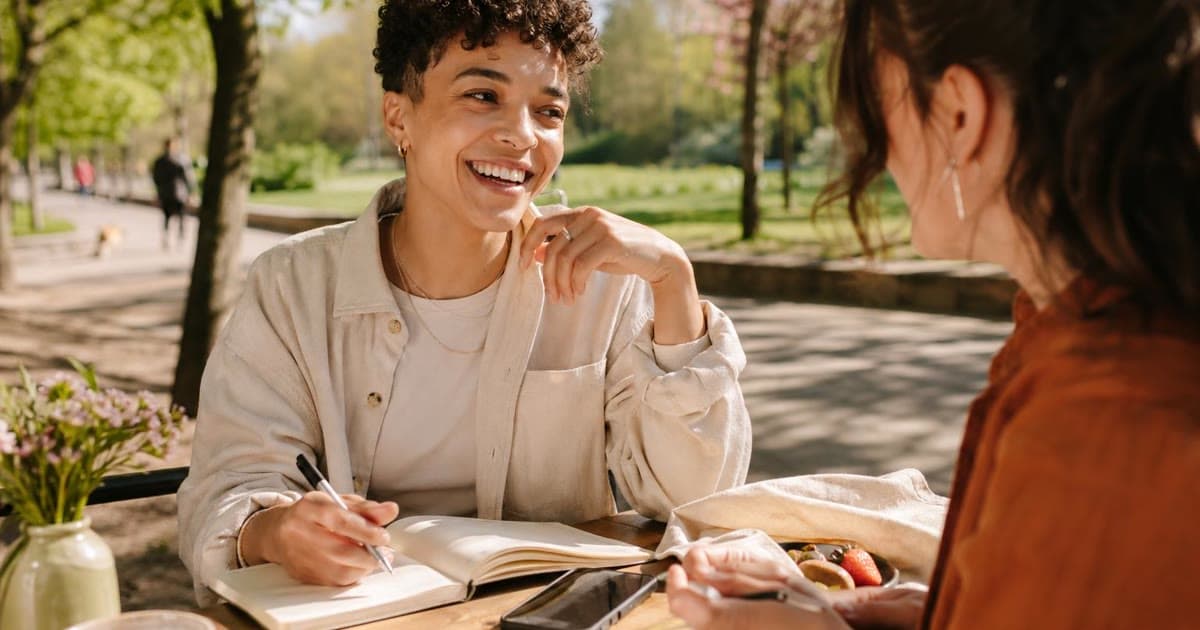 Two people sit at an outdoor table while one holds a pen over an open notebook, with a black phone beside the pages.