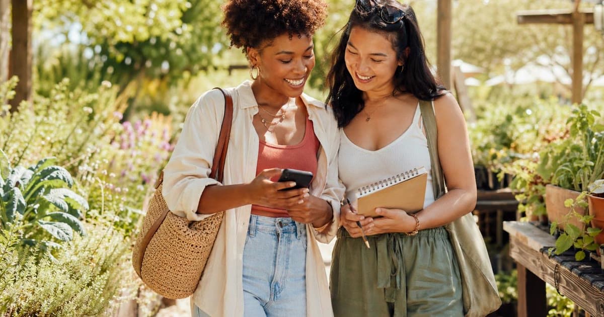 Two young adults stand in a sunny garden looking at a phone while one carries a notebook and pencil.
