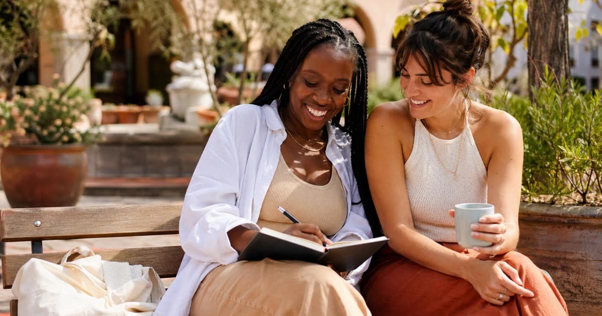 Two women sit together on an outdoor bench while one writes in an open black notebook and the other holds a mug.