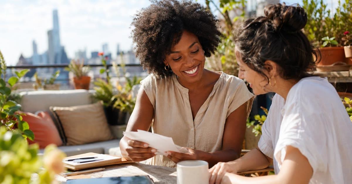 Two people reading a paper together at a sunny outdoor table with a notebook and phone beside them