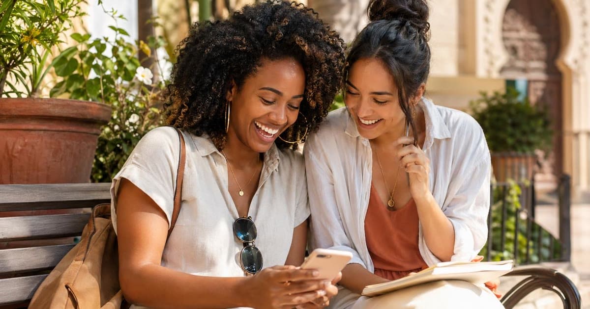 Two women sit on a bench looking at a phone with an open notebook beside them.