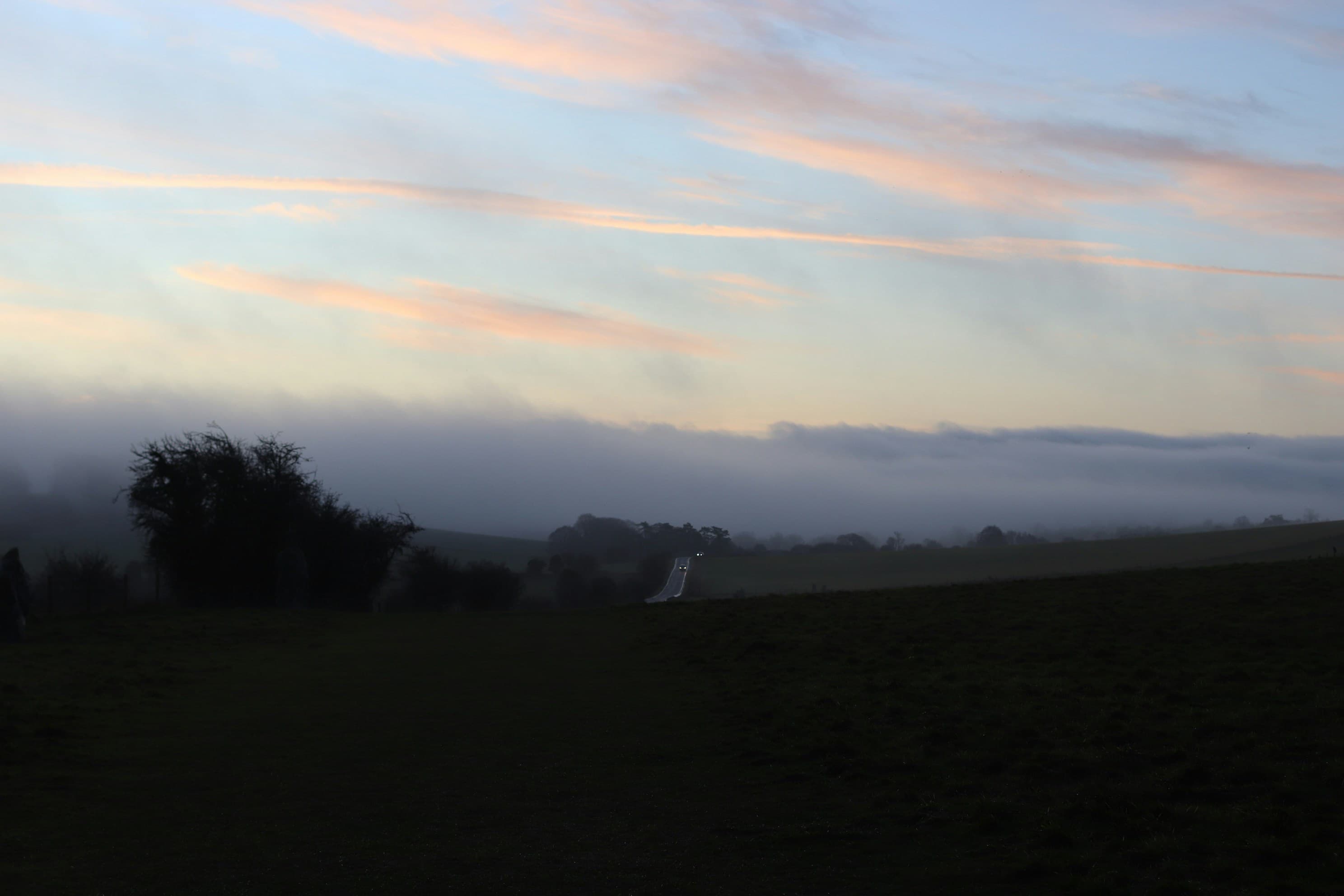 Dark country fields beneath a pale early-morning sky, with a narrow road and distant car lights.