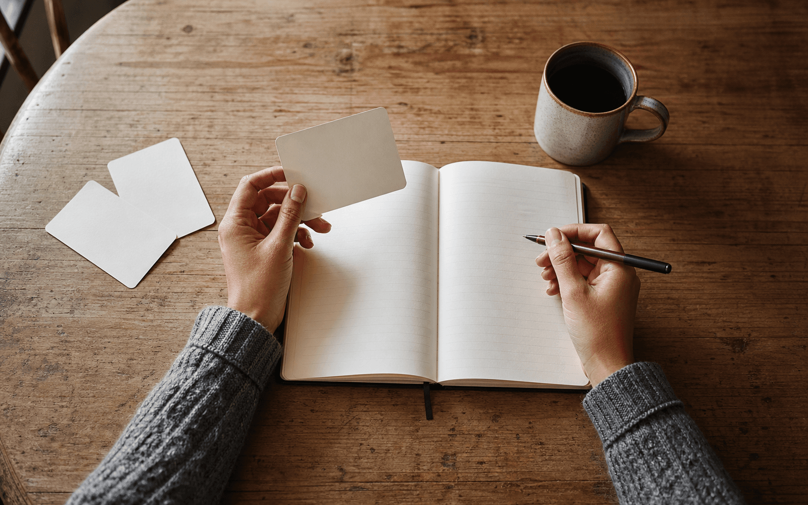 Hands hold a blank prompt card above an open notebook, with three more cards and coffee on a wooden table.