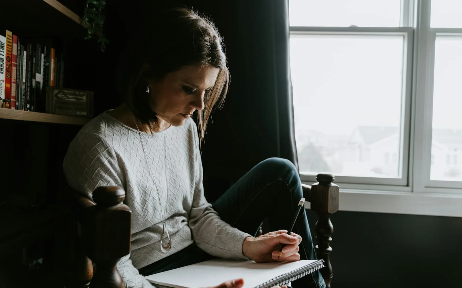 A woman resumes writing in a notebook beside a window.