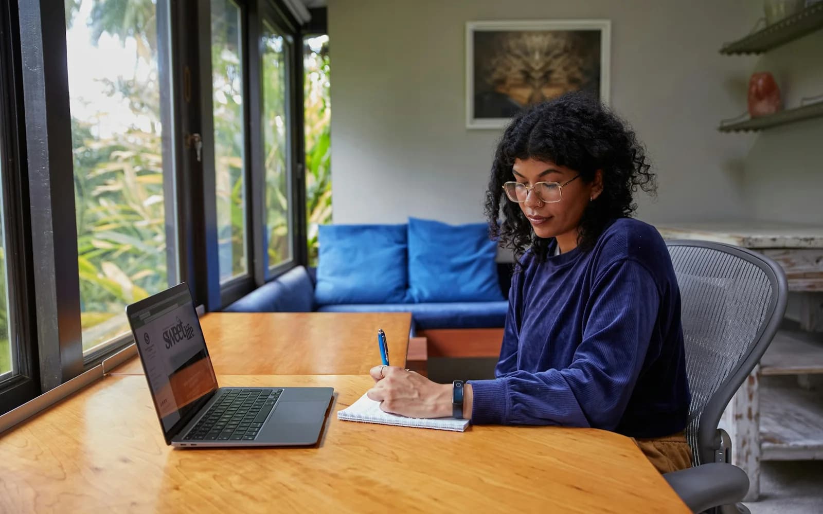 A woman writes notes beside an open laptop at a sunlit table.