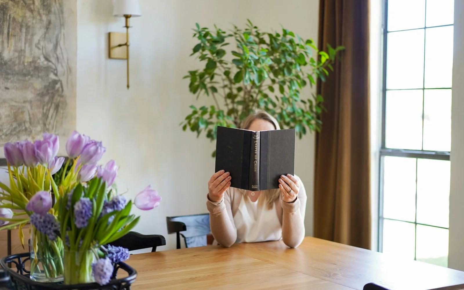 A woman quietly reads a notebook at a dining table.