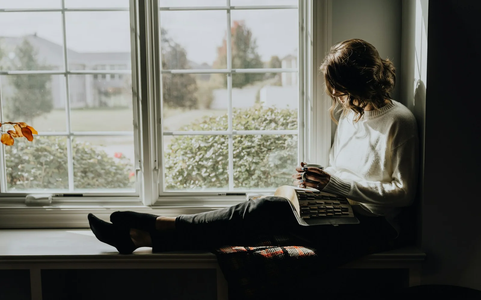 A woman rereads a notebook while sitting beside a window.