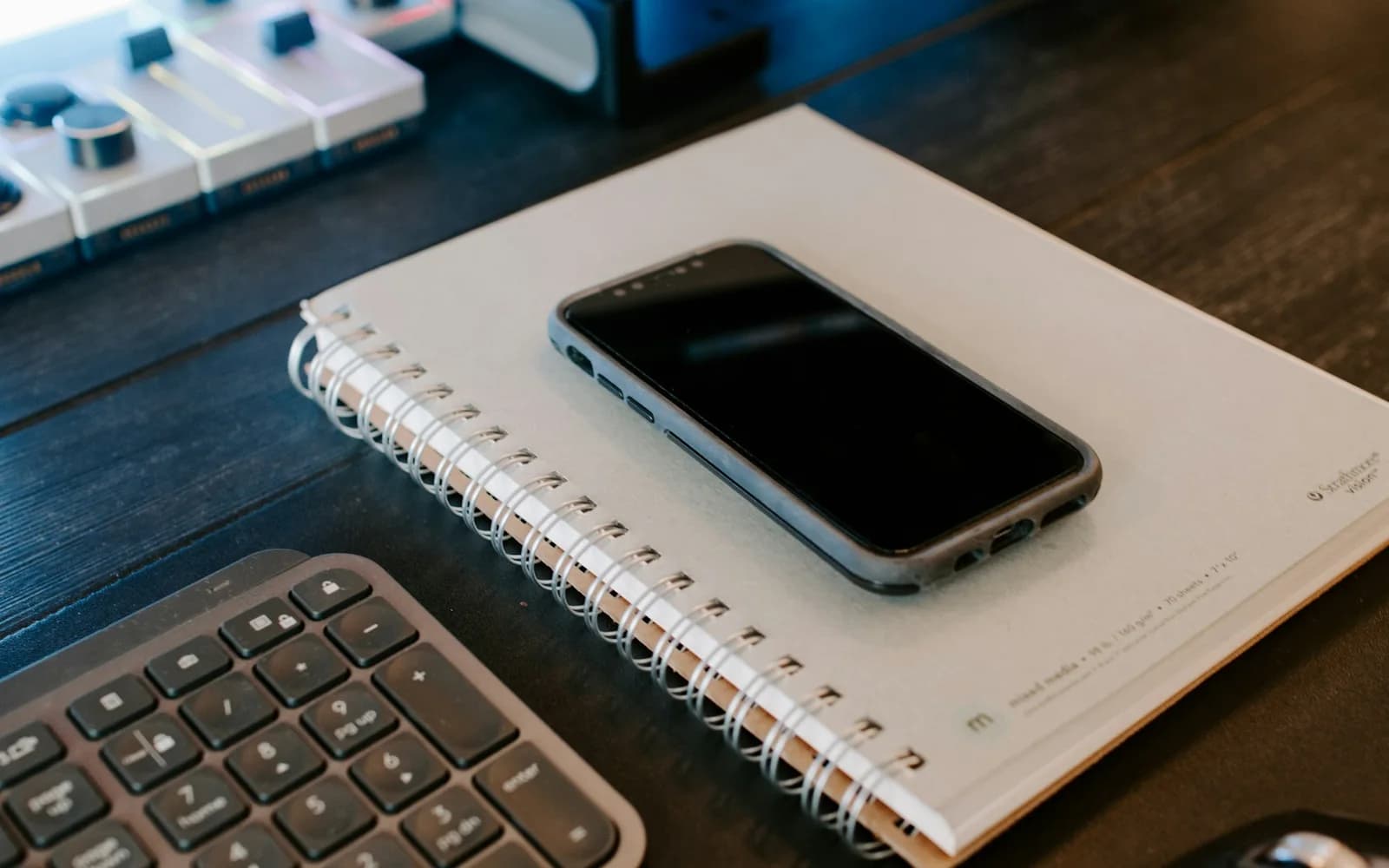 A phone rests on a spiral notebook beside a computer keyboard.