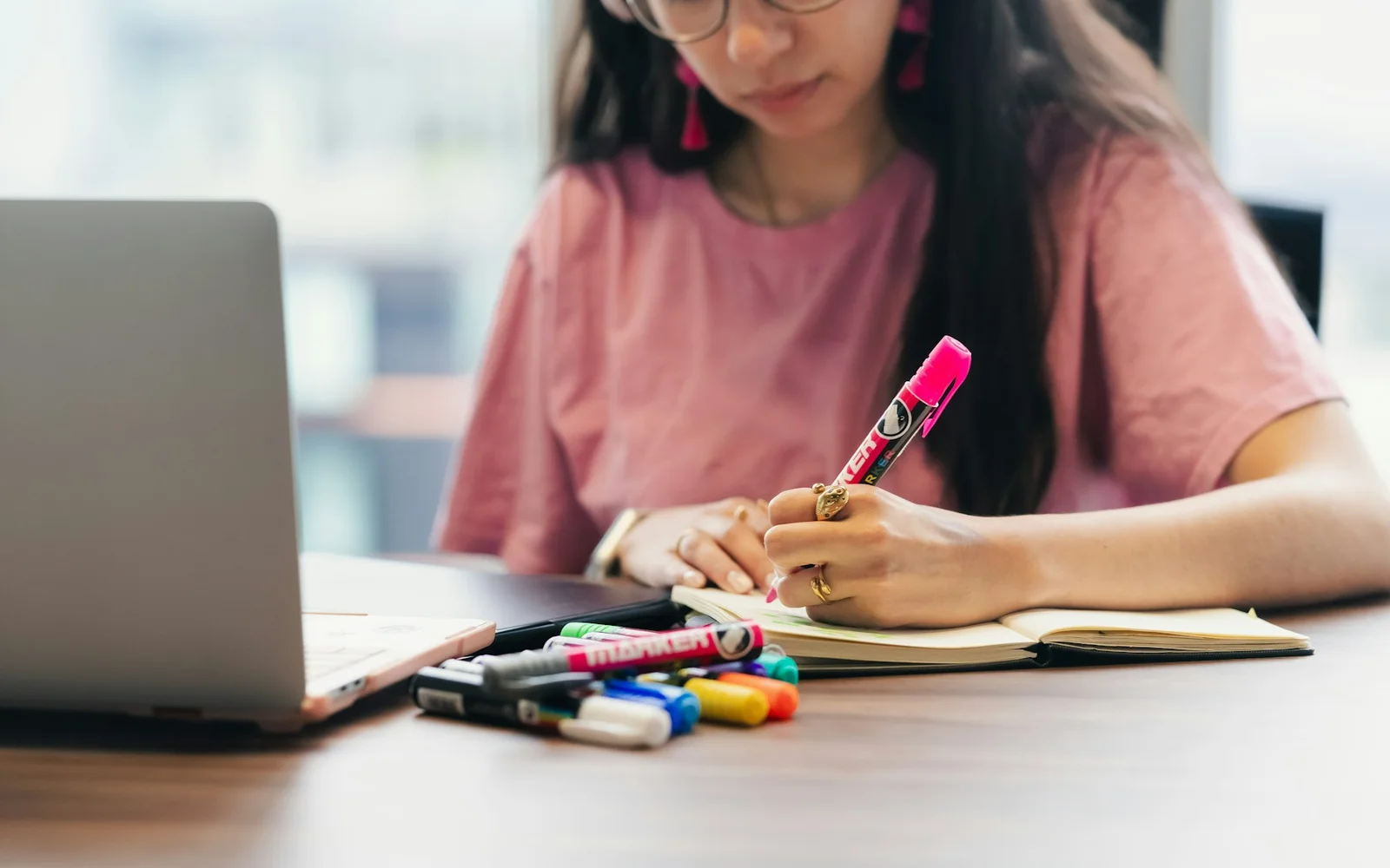 A student writes in a notebook beside a laptop and colored pens.