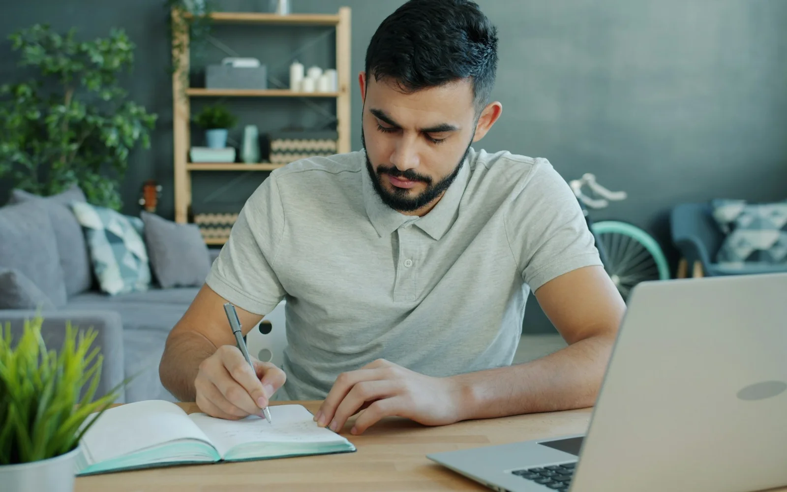 A man writes in a notebook beside his laptop at home.
