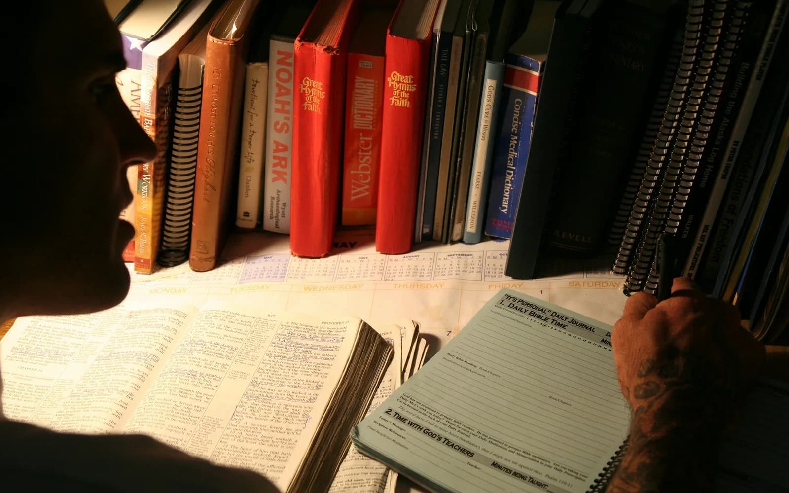 A student writes at a library table surrounded by books.