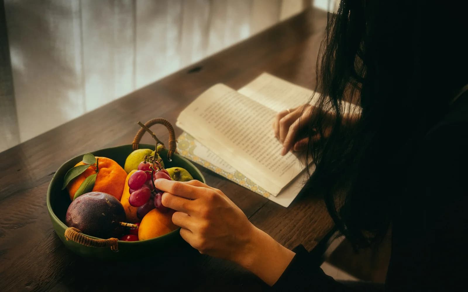 A person reads and writes beside a bowl of fruit in window light.