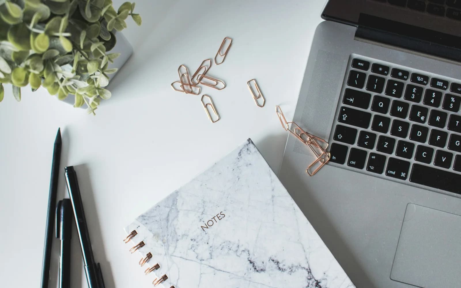 A laptop, notebook, pens, and paper clips arranged on a working desk.