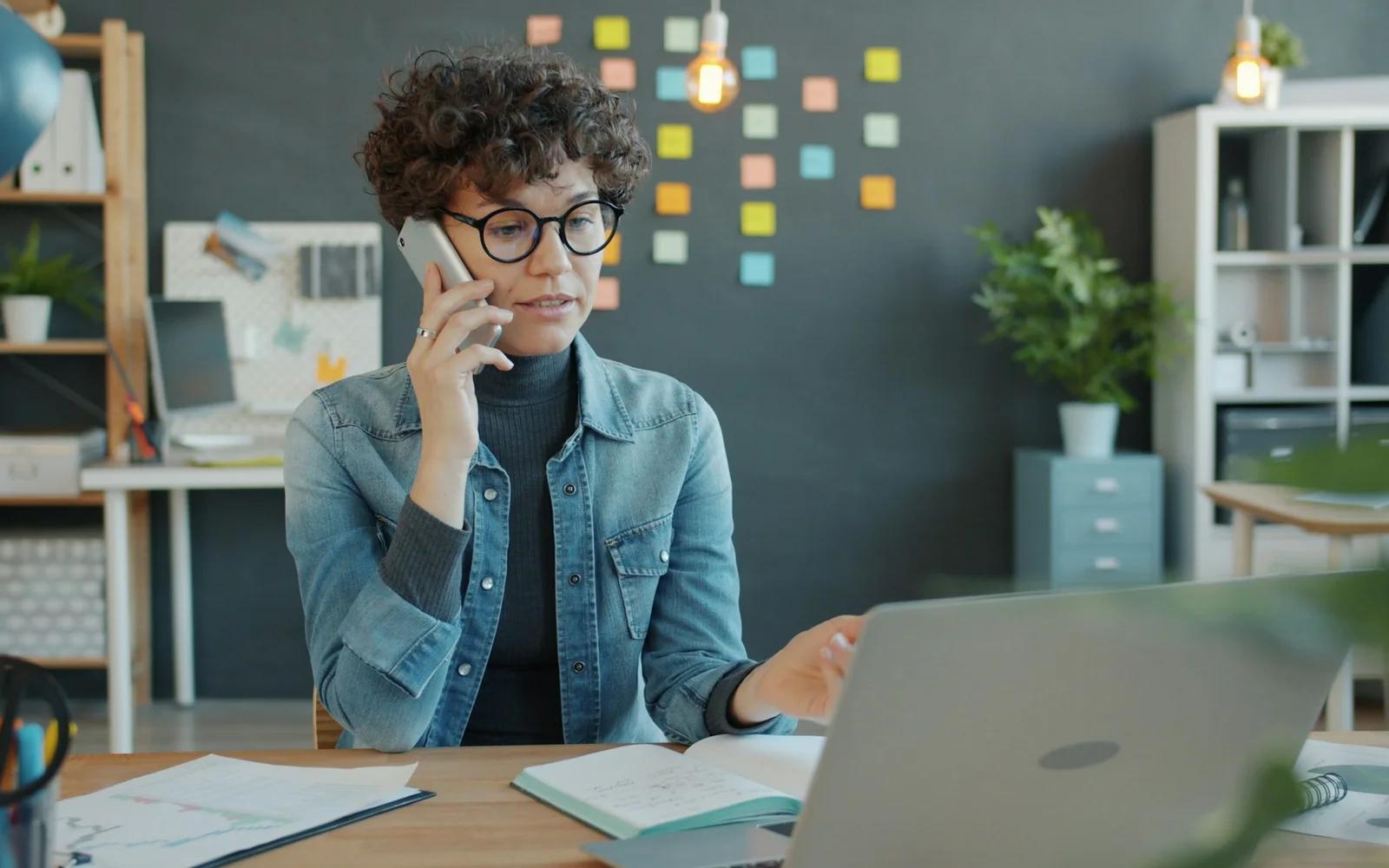 A woman talks on a phone while working beside a laptop.