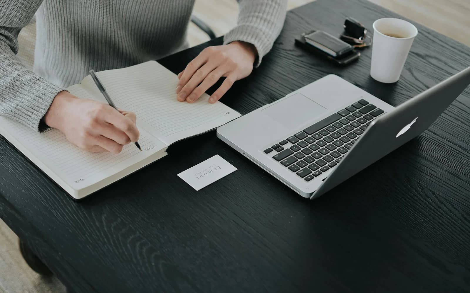 A person writes in a notebook beside a laptop at a home desk.