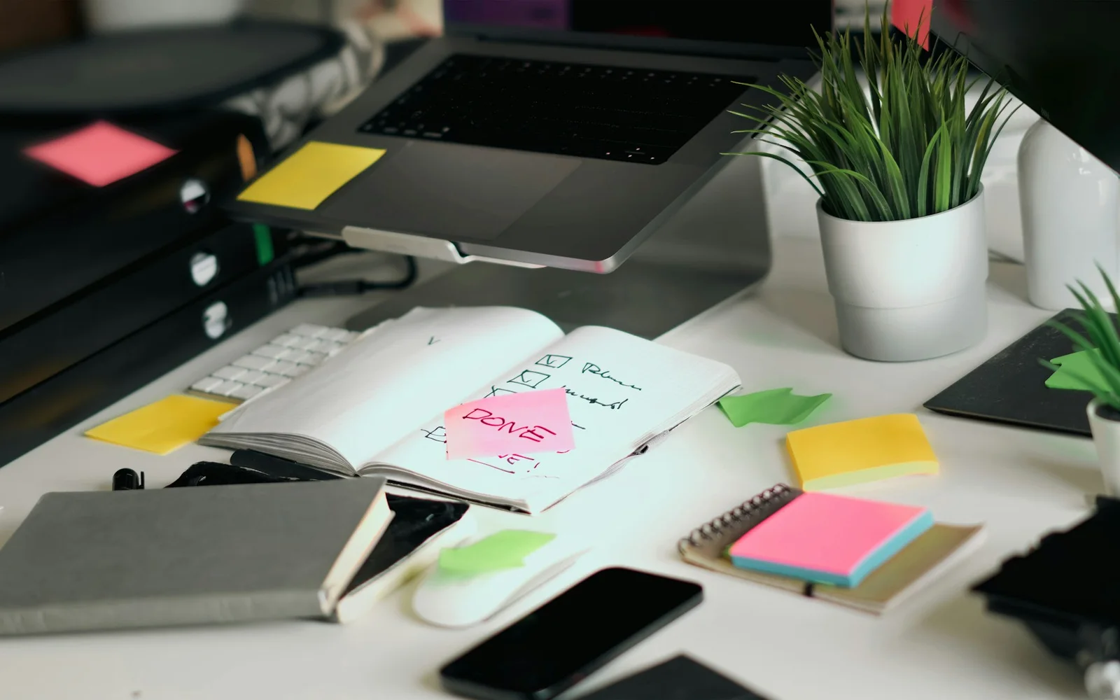 A lived-in desk holds a laptop, notebooks, sticky notes, and pens.