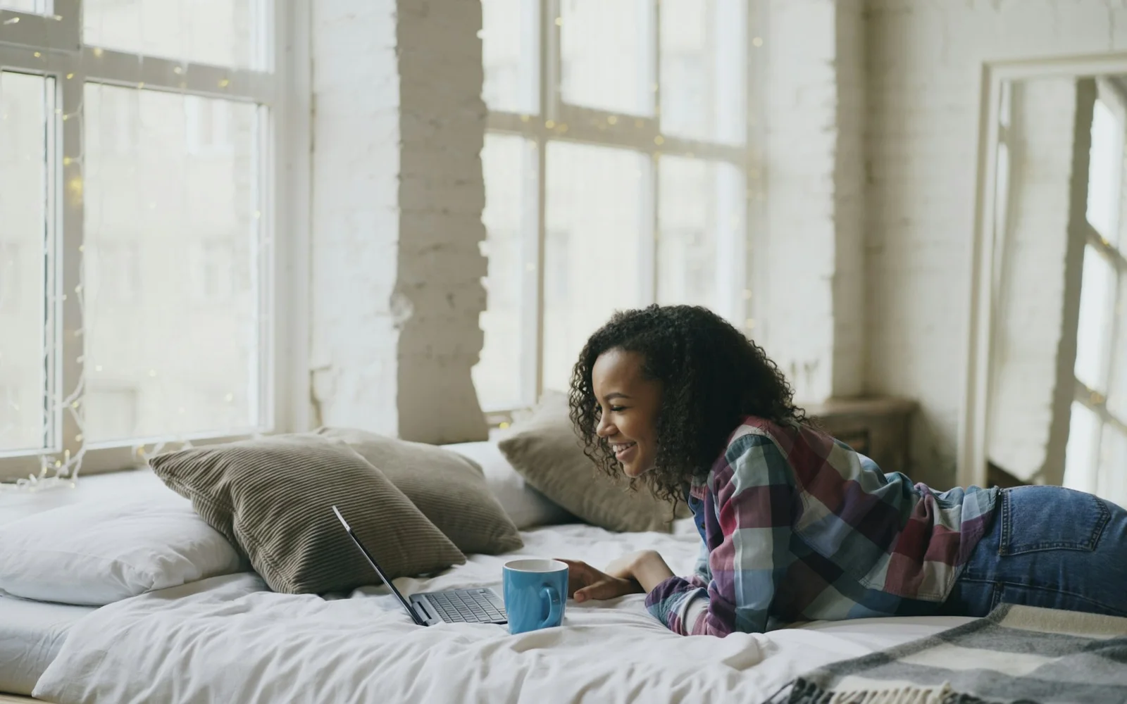 A woman uses a laptop from bed with a notebook and coffee nearby.