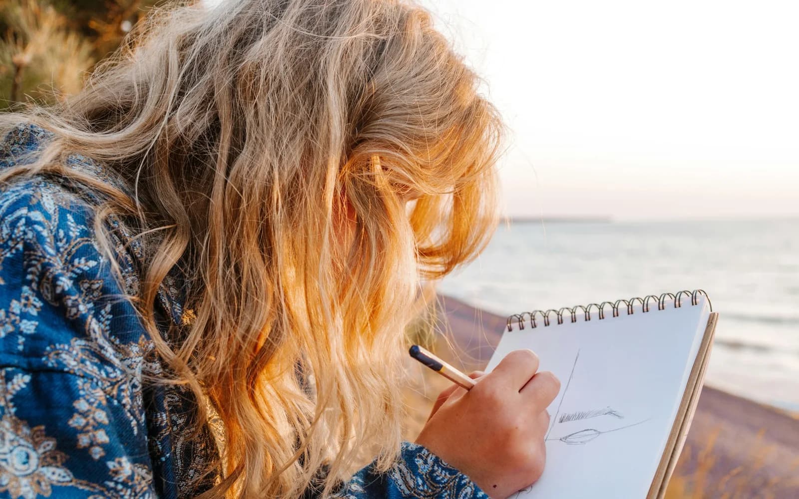 A woman writes in a notebook outdoors beside the water.
