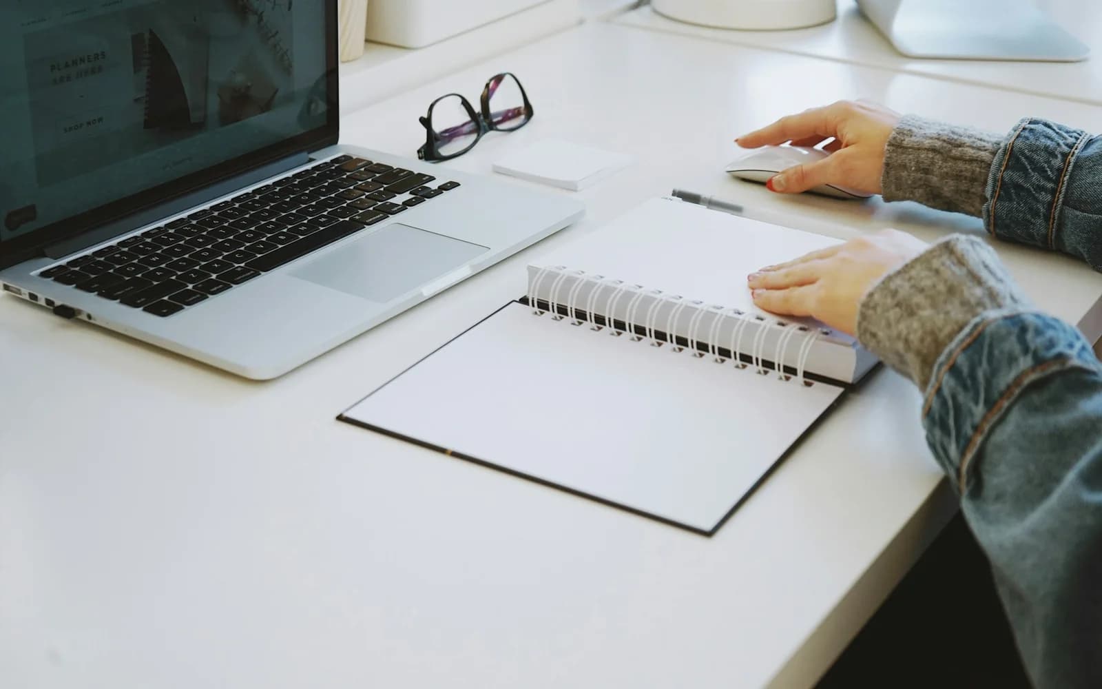 A person turns a spiral notebook beside an open laptop.